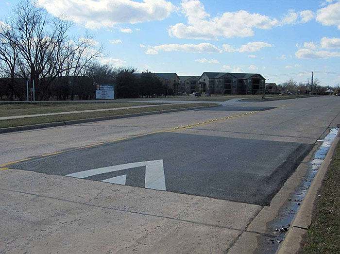 ""Figure 3.13.1. Offset Speed Table. This figure contains a photograph of a straight two lane road divided by a double yellow line. In the near distance in the right lane only, there is a speed table with striping indicating the right of way. A car length further away, there is an offset speed table in the left hand lane. Raised reflectors separate the lanes in between the two speed tables. Off of the left hand side of the street there are apartments in the distance.