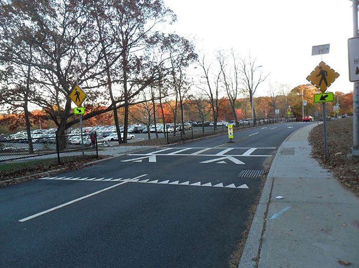 ""Figure 3.14.3. Raised Crosswalk Midblock. This figure contains a photograph of a raised crosswalk on a slightly curved two lane road divided by dashed lines. A sidewalk follows the road on its right side. A signpost holding a diamond shaped yellow sign with icon of a person walking and a fluorescent green rectangular sign with a black arrow pointing downwards at a slight angle is to the right of the sidewalk. The raised crosswalk, with painted arrows indicating right of way and a white ladder shape indicating where to cross, connects the sidewalk and a full parking lot. A sidewalk curves away from the left hand side of the crosswalk and down. Fencing and a strand of thin trees separate the parking lot from the road.