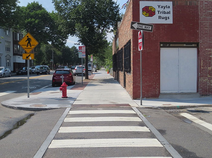 "Figure 3.14.4. Raised Crosswalk at Intersection. This figure contains a photograph of a raised crosswalk at an intersection taken from the point of view of a pedestrian. The white striped pattern crosses to a sidewalk which stretches up the center of the photo. To the right of the sidewalk, there is a red brick single story building. A signpost to the right of the crosswalk just at the curb holds a Do Not Enter sign and a One Way sign pointing to the left. Across the sidewalk from that there is a red fire hydrant and a crosswalk sign. Trees and business line the street to the left of the sidewalk."