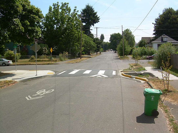 ""Figure 3.14.5. Raised Crosswalk with Curb Extension. This figure contains a photograph of a tree-lined residential area. The focus is a road which runs from the bottom middle of the photo up to the top middle. The road narrows at approximately the halfway point using a curb extension. There is a raised crosswalk that leads from the curb extension to the opposite corner of a sidewalk. Just above the crosswalk, there is an intersection.