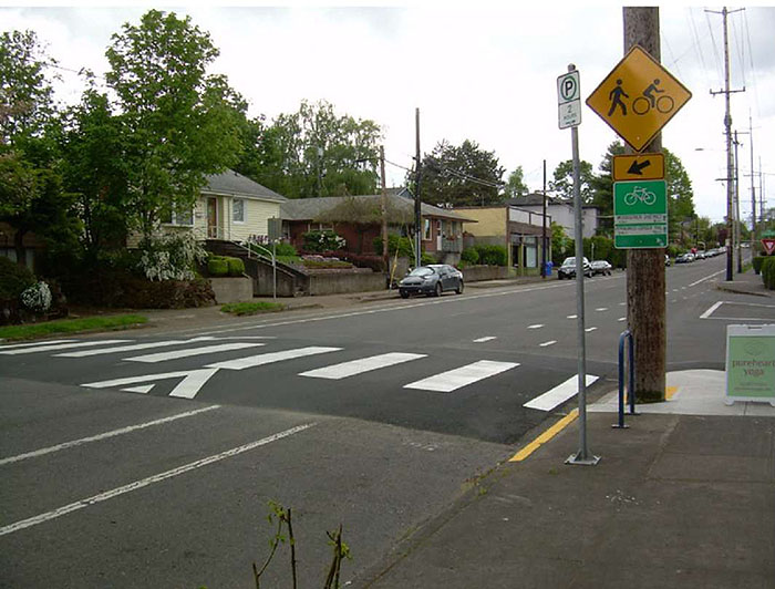 "Figure 3.14.6. Raised Crosswalk with Bicycle Lane. his figure contains a photograph of a city street which runs diagonally from bottom left to top right of the photo. To the right of the right lane, there is a bicycle lane marked with solid white lines and space for parallel parking near the curb. To the right of the crosswalk there is a phone pole holding a diamond sign with the icon of a person walking and a bicycle. There is a rectangular sign below that with an arrow pointing slightly down and to the left. Below that there is a green rectangular sign with a bicycle on it. Across the raised crosswalk, there are trees and a row of houses and other buildings."