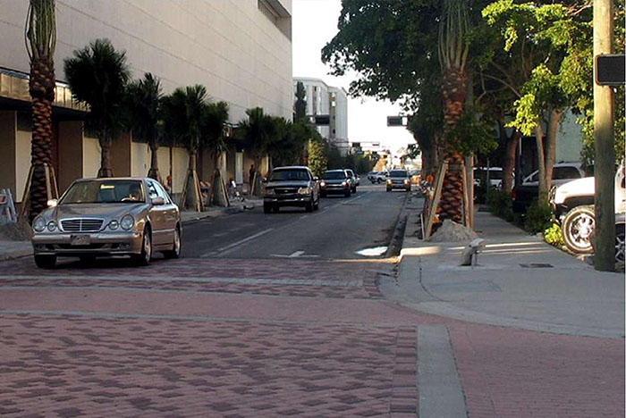 ""Figure 3.15.2. Raised Intersection in Dense Urban Setting. This figure contains a picture of a two lane, one way road between two strands of palm trees and multi-story buildings. There is oncoming traffic in both lanes. Two entrances to the raised intersection can be seen. The intersection itself is done in light red brick.