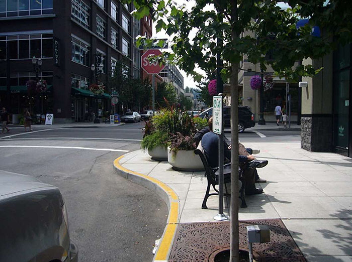 "Figure 3.16.3. Corner Extension in Urban Setting. This figure contains a photograph with a focus on the corner extension. Near the bottom of the picture there is a tree planted at the curb. Further up the sidewalk on the corner extension there is a bench with people seated on it and a series of large, round planters. There is a stop sign at the intersection facing the camera. A four story building occupies the corner diagonally from the extension. The opposite corner has a set of shops."