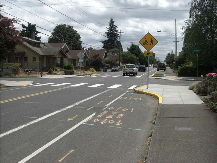 "Figure 3.16.5. Corner Extension and Bicycle Lane. This figure contains a photograph of a street running from the bottom left to the top right. Another street forms an intersection running across the photo's centerline. A signpost can be seen on the corner extension extending into the street running top to bottom. A diamond shaped yellow sign depicts an icon of a person walking and a bicycle. Near the curb there is space for parallel parking and between the parking and the lane there is a separate bicycle lane. Crosswalks are visible running left to right at the intersection. A row of houses can be seen on the corner opposite."