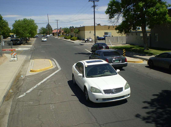 "Figure 3.17.5. Choker with Passing Traffic. his figure contains a photograph of an undivided two lane street running from the bottom right to near the top left of the picture. Curb extensions, detached from the sidewalks and outlined in white stripes, dominate the center of the photograph. A white car travelling towards the camera and a gray car travelling away are passing through the choker at the same time.  A car is parallel parked in the opposite lane, pointed away from the camera after the chocker. Trees are scattered around the sides of the street and a  tan building can be seen in the distance."