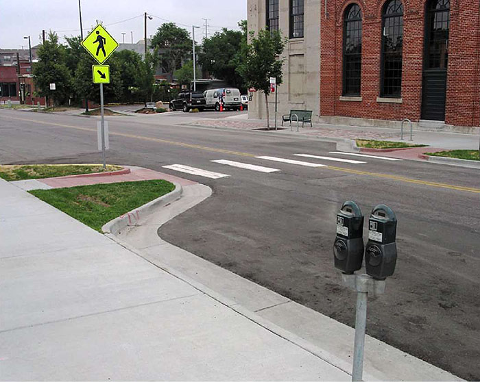 "Figure 3.17.7. Choker Adjacent to On-Street Parking. This figure contains a photograph of a two lane street divided by a double yellow line, using a choker at a crosswalk. The left hand side of the street shows available parallel parking near two parking meters. Buildings and off street parking can be seen across the street. The curb extensions are landscaped with grass on either side of the ramps to the sidewalk."