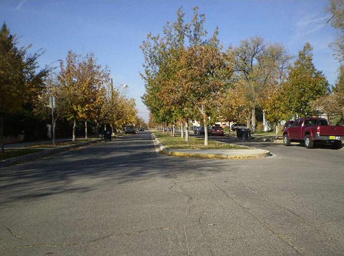 "Figure 3.18.9. Long Midblock Median Island. This figure contains a photograph of a two lane road separated for its visible distance by a wide, long median island. The island is landscaped along its length with grass and a single line of trees. Traffic, including a red pickup truck, can be seen in the right hand lane. More trees line either side of the street."