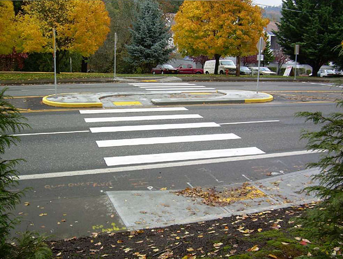 "Figure 3.18.10. Median Island with Pedestrian Refuge and Offset Crosswalk. This figure contains a photograph of a four lane road running from left to right. The section of crosswalk traversing the nearest two lanes leads to a large, oval island with a recessed sidewalk. The walkway curves to the right along the island's length and the crosswalk resumes, offset to the right. A small parking area can be seen across the street along with a number of trees."