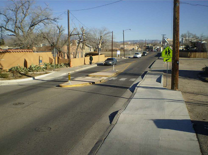 "Figure 3.18.11. Median Island with Pedestrian Refuge. his figure contains a photograph of a two lane street. A median island functions as a place for anyone using the crosswalk to wait for oncoming traffic to pass. The right hand side of the street appears to be a parking lot. A pedestrian crossing sign points at the crosswalk. On the left hand side of the street there is a small residential area."