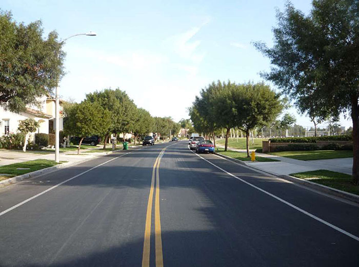 "Figure 3.19.1. On-Street Parking Near Residences and Park. This figure contains a photograph of a two lane road divided by a double yellow line. The street runs from bottom center to top center of the picture. Between the lane and curb on either side, areas for parallel parking are delineated by solid white lines. The right hand side of the street is tree line next to a park. The left hand side of the street is also tree-lined and several houses can be seen."