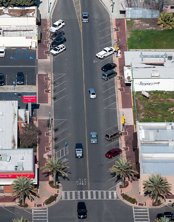 "Figure 3.19.2. On-Street Parking in a Commercial Business District. This figure contains a photograph of a two lane street divided by double yellow lines. The picture was taken from high overhead. Parking spots on either side are diagonal relative to the sidewalk. Businesses line both sides of the street. There a several slots open on both sides and light traffic on the road. A crosswalk and corner extensions can be seen near the bottom of the picture."