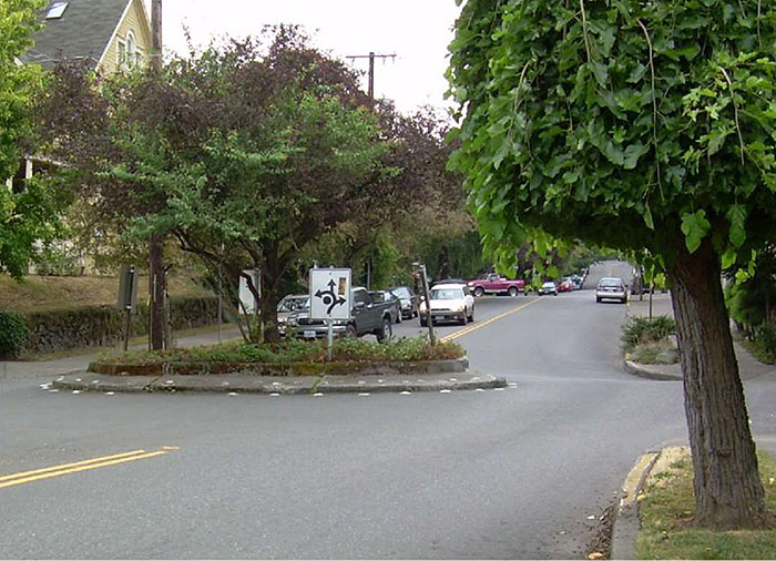 Figure 1.1. Horizontal Deflection Measure - Traffic Circle. Please see the Extended Text Description below. This figure contains a photograph of a traffic circle as a horizontal deflection measure. The center of the traffic circle contains trees, shrubs and a sign showing a curved line with three arrows coming off of it, indicating the exits from the circle. Oncoming traffic includes a gray truck and a white car