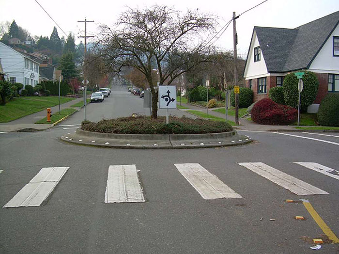 Figure 3.7.5. Potential Pedestrian-Vehicle Conflict at Traffic Circle. This figure contains a photograph of a traffic circle in a residential neighborhood. The concrete circle has white dots following its circumference. The center contains one tree and groundcover. White, square signs facing each section of street show that traffic should keep right and the points at which the circle can be exited using curved lines and arrows. In the foreground near the traffic circle, there is a crosswalk.