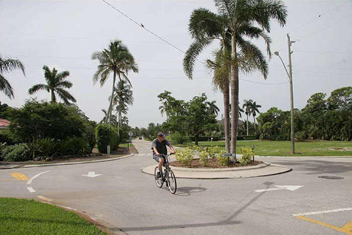 Figure 3.7.6. Bicyclist Passing Through Traffic Circle. This figure contains a photograph of a traffic circle in a residential neighborhood. The concrete circle contains palm trees and low shrubs. White, square signs facing each section of street and white arrows painted on the street surface show that traffic should keep right. A person on a bicycle is riding through the traffic circle, approaching the section of street near the photo's bottom right corner.