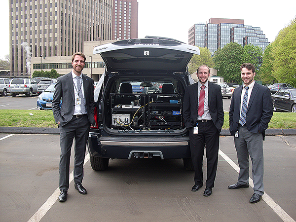 "Photograph. Instrumented research vehicle (IRV) used to collect data through work zones with Taylor Lochrane (left) from FHWA, Christopher Melson (center) from FHWA, and Andrew Berthaume (right) from the Volpe Center. The back of a blue 2007 Jeep Grand Cherokee is open, showing electronic data collection equipment installed on black metal brackets. Three young gentlemen in suites and ties pose around the Jeep. The Jeep is stationed in a parking lot, in an urban setting with surrounding brick buildings."