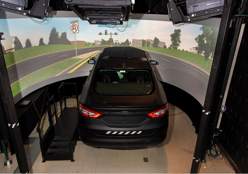 "The highway driving simulator comprising of a large, curved projection screen, showing a simulated road and intersection, in front of a car connected to the simulation computers."