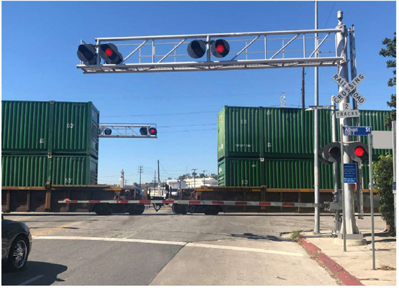 "Figure 21. Illustrative Example Showing Cantilever with Flashing-Light Devices - This figure shows a photograph of a fundamental active warning device in use as a train is crossing and a car is stopped before the crossing. It is kept a safe distance from the train. The fundamental active warning device in the photo is a flashing-light signal, installed on cantilevered supports for additional emphasis and better visibility for approaching traffic. Installing a flashing-light device on an overhead structure or cantilevered supports as shown in the photo provides better visibility for multi-lane approaches to railroads or highways with profile restrictions."