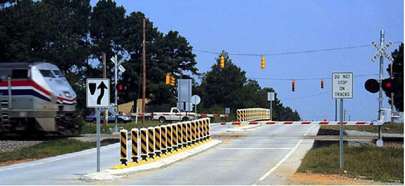 "Figure 37. Example of a Traffic Separator - This figure is a photograph showing an example of a mountable raised curb system, also known as a "traffic separator." In the photograph, a train is approaching from the left. To the right of the traffic separator, there is a Crossing Gate, which is attached to the Crossing Gate. There is an additional Crossing Gate with a traffic separator attached to it on the other side of the street. It is facing the roadway for oncoming traffic. There are several flexible tubular delineators (aka vertical panels) on both traffic separators."