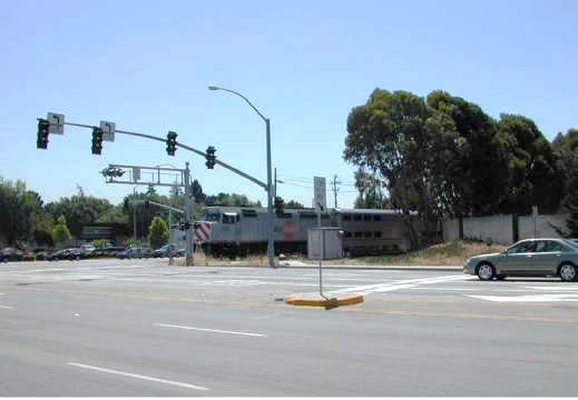 "Figure 40. Illustrative Example Showing Displaced Stop Line (PCJX S. Mary Avenue Crossing at W. Evelyn Avenue, Sunnyvale, CA, USDOT 755037b) - This figure is a photograph of a vehicle a safe distance away from an approaching train about to cross through an intersection in front of the driver. Despite the long distance to the railroad crossing and the intersection, the driver is complying with the STOP HERE ON RED signal."