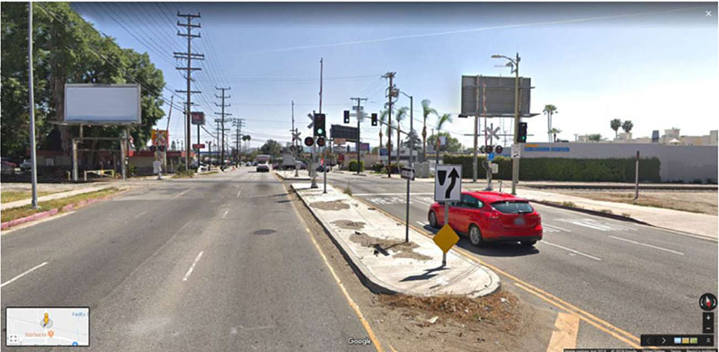 "Figure 43. Illustrative Example of Queue Cutter (SCAX Balboa Boulevard Crossing at Roscoe Boulevard in Los Angeles, CA, USDOT 745989G) - This figure is a photograph of a red car about to go through a greenlight crossing over an intersecting railroad on two-way, 3 lane street. This picture shows a queue cutter, which utilizes detection loops. There is also a mountable raised curb system on both sides of the railroad."