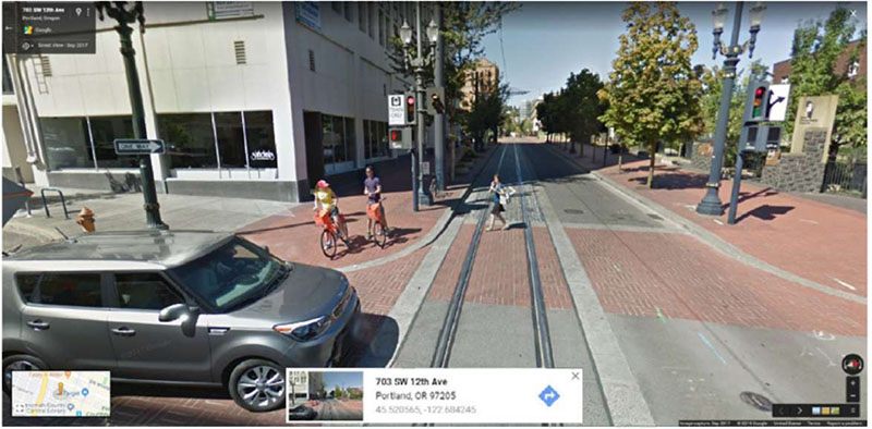 "Figure 50. Illustrative Example of Dynamic Envelope Delineation through a Pedestrian Crosswalk (Portland TriMet, Morrison Street, Portland, Oregon) - This figure is a photograph of a car about to cross over an LRT trackway that pedestrians are also crossing. There is no train present, so we can see the contrasting pavement to delineate the dynamic envelope through a crosswalk."