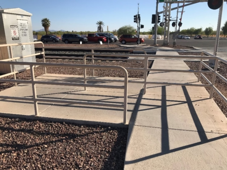 "Figure 52. Illustrative Example of a Pedestrian Barrier Application (BNSF Cotton Crossing. Peoria, AZ USDOT 025405Y) - This figure is a photograph of a pedestrian barrier application near a sidewalk crossing at a railroad. It appears to be in an offset pattern, creating a "maze" to force pedestrians to turn and look both ways approaching a crossing specifically for pedestrians. There is a contrasting pavement color and texture from the railroad."