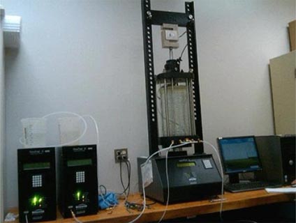 Figure 3. This photo shows a sample being tested in a large 6-inch diameter triaxial chamber within a load testing device equipped with a load cell. The device is connected to two water pumps through clear plastic tubes and controlled with a data collection computer.