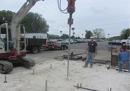 Figure 13. The image shows two men working in a sand pit. To the left of the pit is a piece of heavy equipment that is drilling steel anchors into the pit.