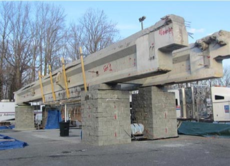 Figure 16. This photo shows four GRS piers with concrete masonry units (CMUs) on a concrete pad. Two large concrete I-beams are positioned on top of the GRS piers.  The beams have salt spray catch barriers attached to them.