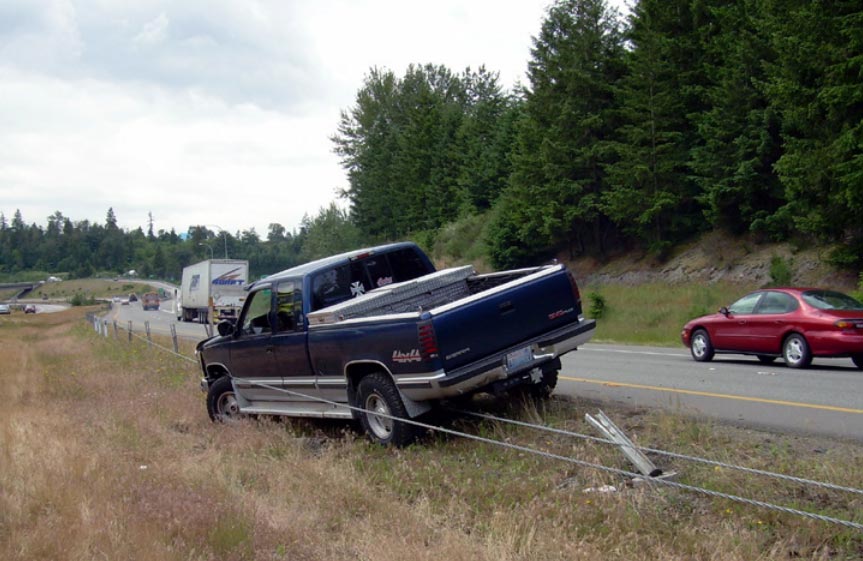 "This photograph, taken from the median of a four-lane divided highway, shows a pickup truck restrained by a median cable barrier. The median cable barrier consists of two horizontal metal cables supported by metal posts. The pickup truck pointing towards opposing traffic at an acute angle. One of the metal cables that comprise the median cable barrier is located underneath the truck, while the other is running along the driver side of the vehicle."