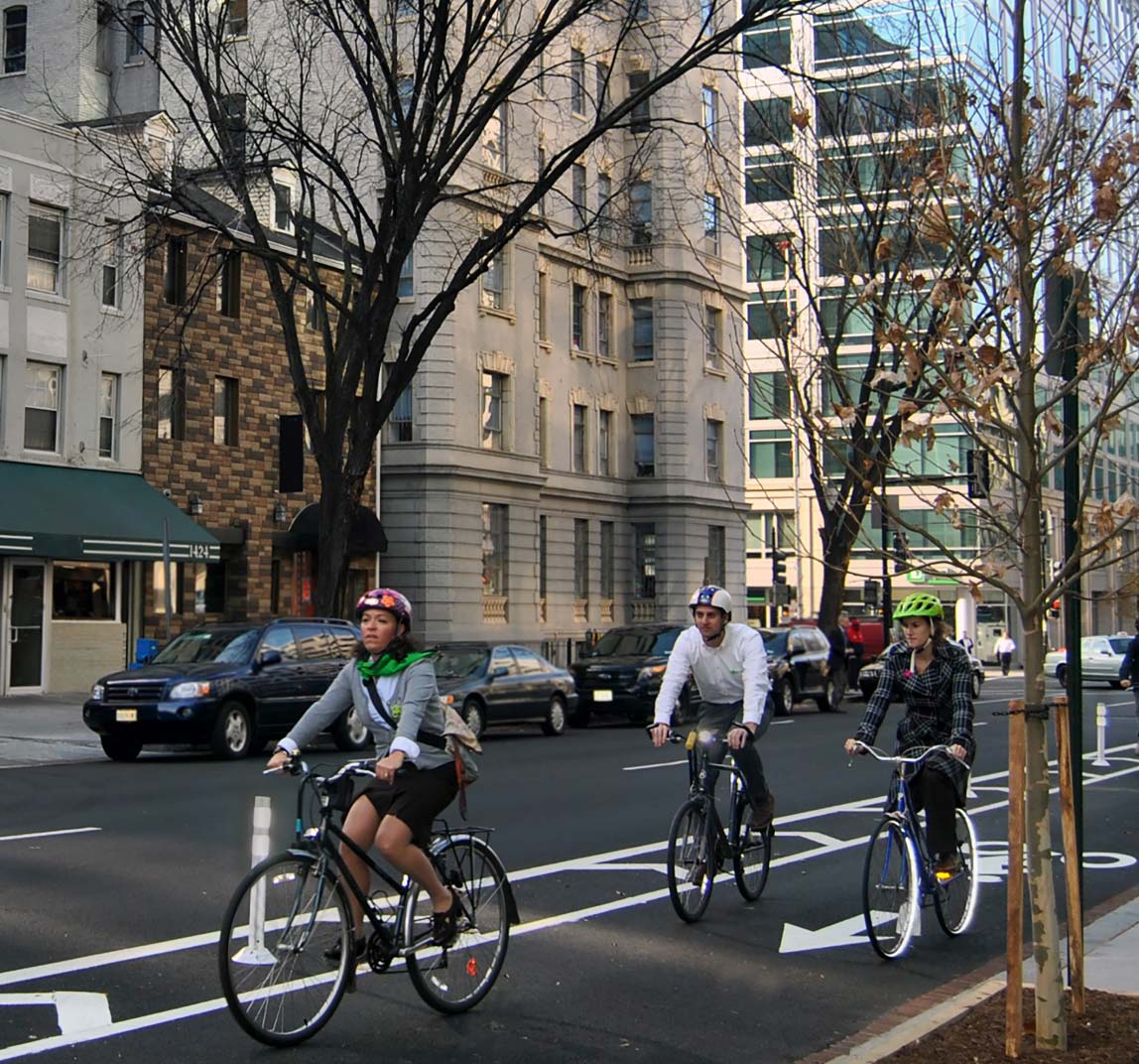 "Photo: This photograph, taken from the edge of the roadway, shows three bicyclists traveling in a buffered bicycle lane toward the foreground of the image in an urban setting. To the left of the bicycle lane are two general travel lanes with on-street parking. The bicycle lane is separated from the roadway with white pavement markings and vertical posts. The bicycle lane is marked with a helmeted bicyclist symbol and directional arrow. To the right of the bicycle lane is concrete curb and sidewalk."