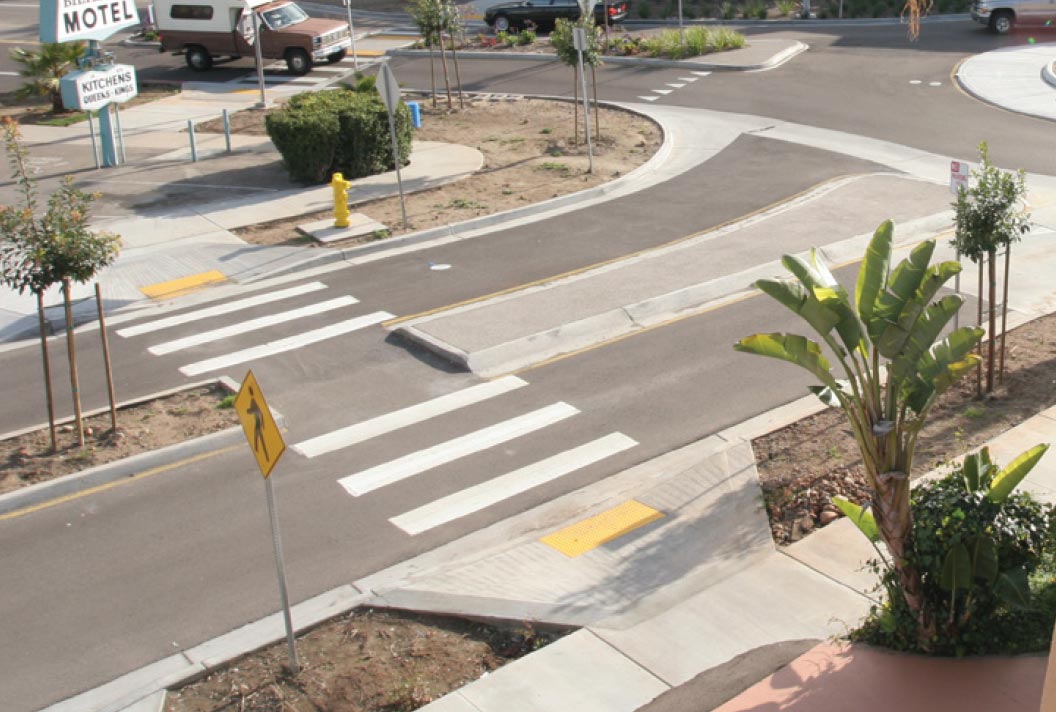 "Photo: This photograph shows a pedestrian refuges island at the approach of a single-lane roundabout. A high visibility crosswalk is located more than a vehicle length away from the roundabout. This crosswalk features pedestrian warning signs, yellow tactile curb ramps, and a cutout within the raised median."