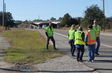 "A group of five individuals inspecting the roadway. The five individuals are standing in the paved shoulder of the roadway and wearing reflective safety vests.