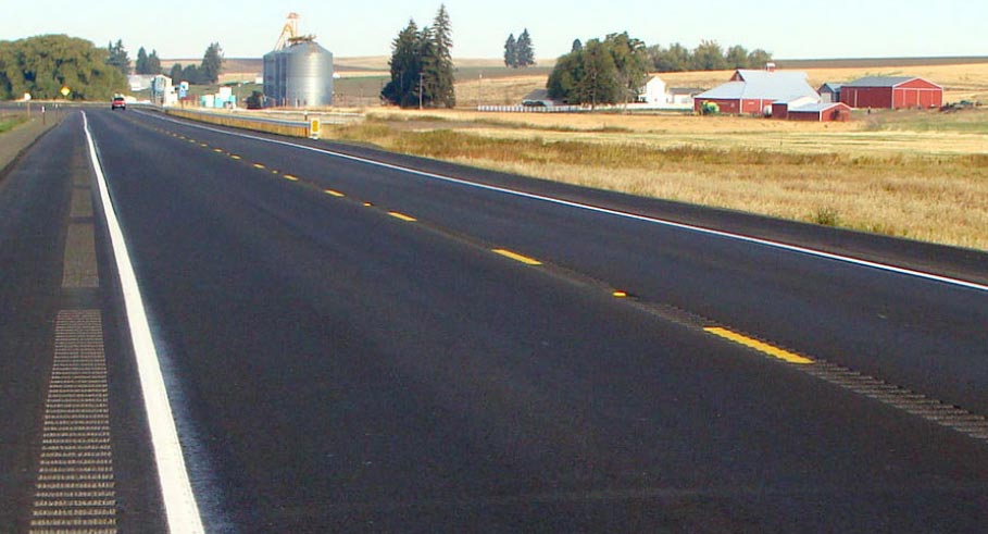 "Photo: This photograph, taken along the edge line of a two-lane undivided roadway, shows longitudinal rumble strips present in the center and along the paved shoulder of the roadway. The roadway is located in a level, rural setting, and the roadway is marked with a dashed yellow centerline and solid white edge lines."