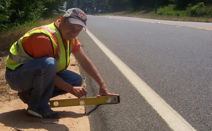 "The angle of SafetyEdge relative to the roadway surface. A person holds a level perpendicular to the edge of the roadway. There is no backfill material adjacent to the paved surface, revealing the SafetyEdge below. The SafetyEdge consists of a slope of asphalt installed at approximately a 30-degree angle to the horizontal."