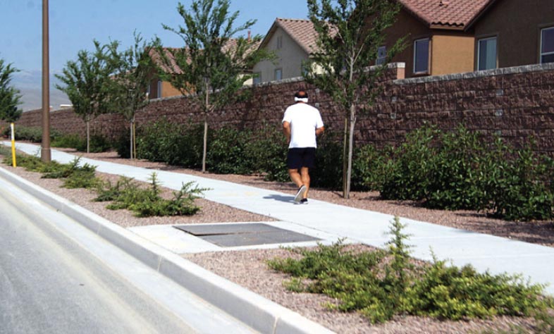 "Photo: This photograph, taken from the roadway, shows a sidewalk in a residential area. The concrete sidewalk is separated from the roadway by a concrete curb and landscaping area. A pedestrian can be seen running on the sidewalk."