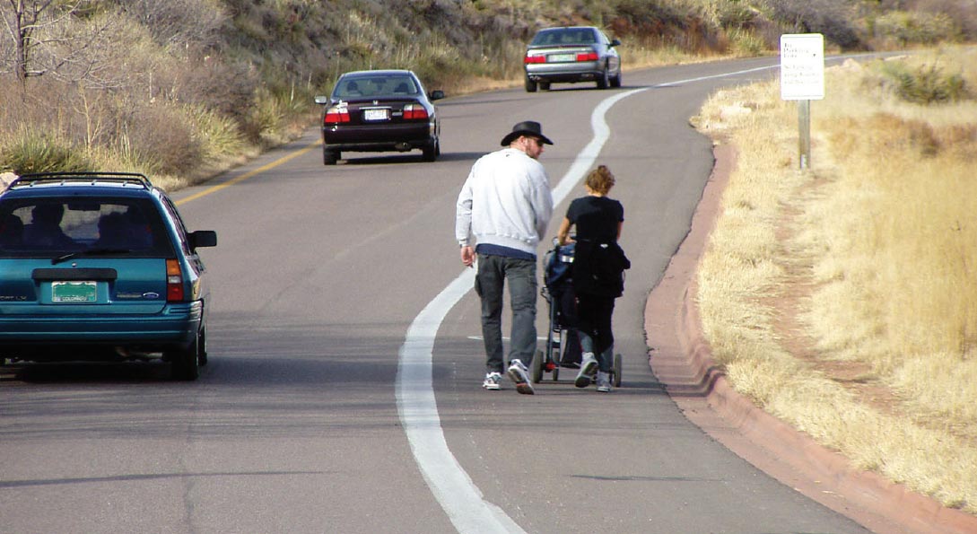 "alt="Photo: This photograph, taken from the roadway, shows a paved shoulder being used as a walkway by two people in a rural area. Vehicles on the roadway are shown traveling in the same direction, indicating that the roadway is a one way or a divided facility. The paved shoulder and general travel lane are separated by a wide, solid white line. The outside of the paved shoulder is bordered by a concrete curb. Beyond the curb, a footpath is visible in the adjacent grass."