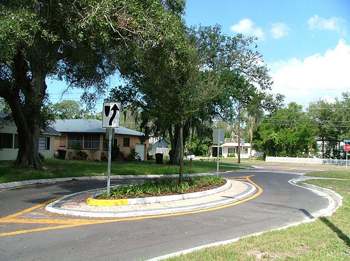 "Figure 3.18.3. Landscaped Oval Median Island. This figure contains a photograph of a street running through a residential area. The only part of the street that's visible is the section using an oval median island to narrow the lanes. The island is landscaped with a tree and low ground cover. A white sign indicates right of way around the island. Several houses are visible across the street from the camera."