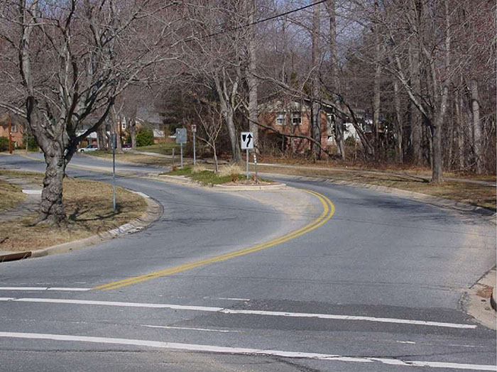 ""Figure 3.18.4. Median Island Near Curve at Entrance to Residential Area. This figure contains a photograph showing the entrance to a residential area. As the road curves to the left, a median island is used to narrow the lanes. The island contains some landscaping and street signs. The only clearly visible sign is the one indicating right of way. Bare trees line either side of the street. Two houses are visible through trees in the distance.