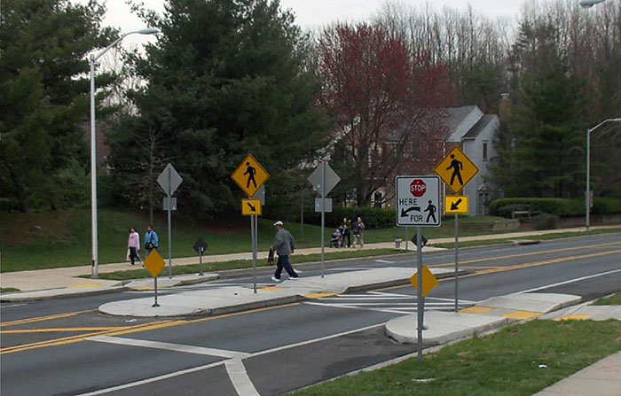 "Figure 3.18.5. Median Island with Crosswalk. This figure contains a photograph of a divided two lane street. Focus is on an island with a crosswalk. Curb extensions are also used to narrow the lanes near this island. A pedestrian is crossing from right to left and is just leaving the island to cross the far lane. Two other pedestrians are walking on the far sidewalk. A white, square sign on the near side of the street shows that cars should stop at the crosswalk for pedestrians. A yellow diamond shaped sign with the icon of a pedestrian and a square sign with an arrow points to the crosswalk. Similar signs occupy the island and far sidewalk."