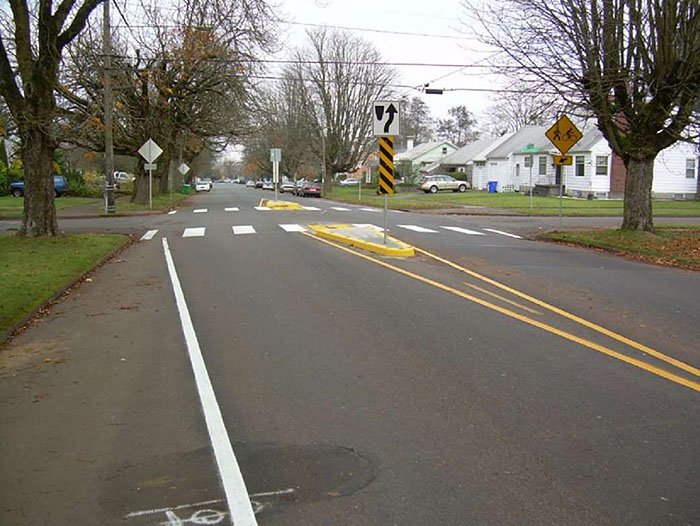 "Figure 3.18.7. Small Median Islands at Intersection. This figure contains a photograph focusing on a road running from bottom to top. An intersection in the top third of the picture shows the use of small median islands before and after the intersection to narrow the lanes. The islands are rounded triangles of concrete containing yellow and black-striped  signs and white signs showing right of way. Large trees line either side of the streets and houses can be seen on the far right side of the street."