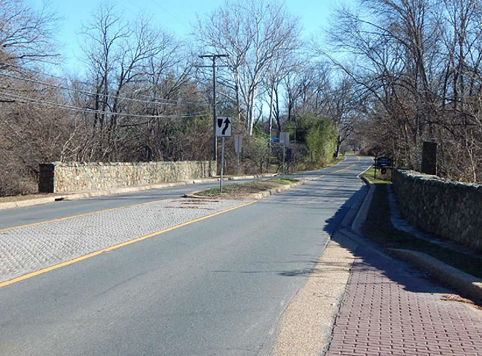 "Figure 3.18.8. Short Midblock Median Island. This figure contains a photograph of a two lane street which uses a median island to narrow the lanes where there are sections of stone walls on either side of it. Trees and heavy undergrowth fill either side of the street beyond the walls."