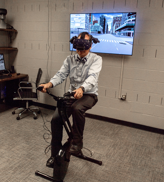 "A volunteer, wearing a virtual reality headset, sits on a stationary bicycle with a monitor on the wall behind him that is showing a simulated intersection.""