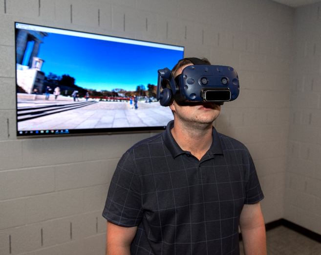 "A volunteer stands, wearing a virtual reality headset, in front of a monitor showing a simulated environment.""