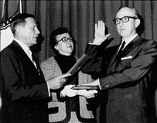 Secretary John A. Volpe administers the oath of office to Director of Public Roads Ralph R. Bartelsmeyer, whose wife Marjorie holds the bible.