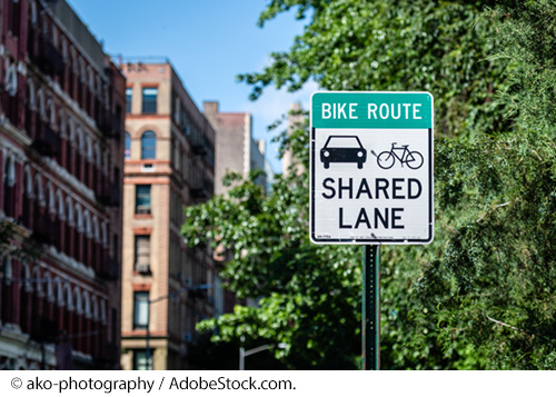 A sign along a city street reads, "Bike Route. Shared Lane." Several buildings and trees can be seen in the background. Image Source: © ako-photography / AdobeStock.com.