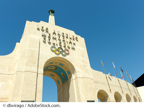 A large entryway leads into a sports arena flanked with various flags to the right of the entrance. Over the entryway reads Los Angeles Memorial Coliseum. Image Source: © dbvirago / AdobeStock.com.