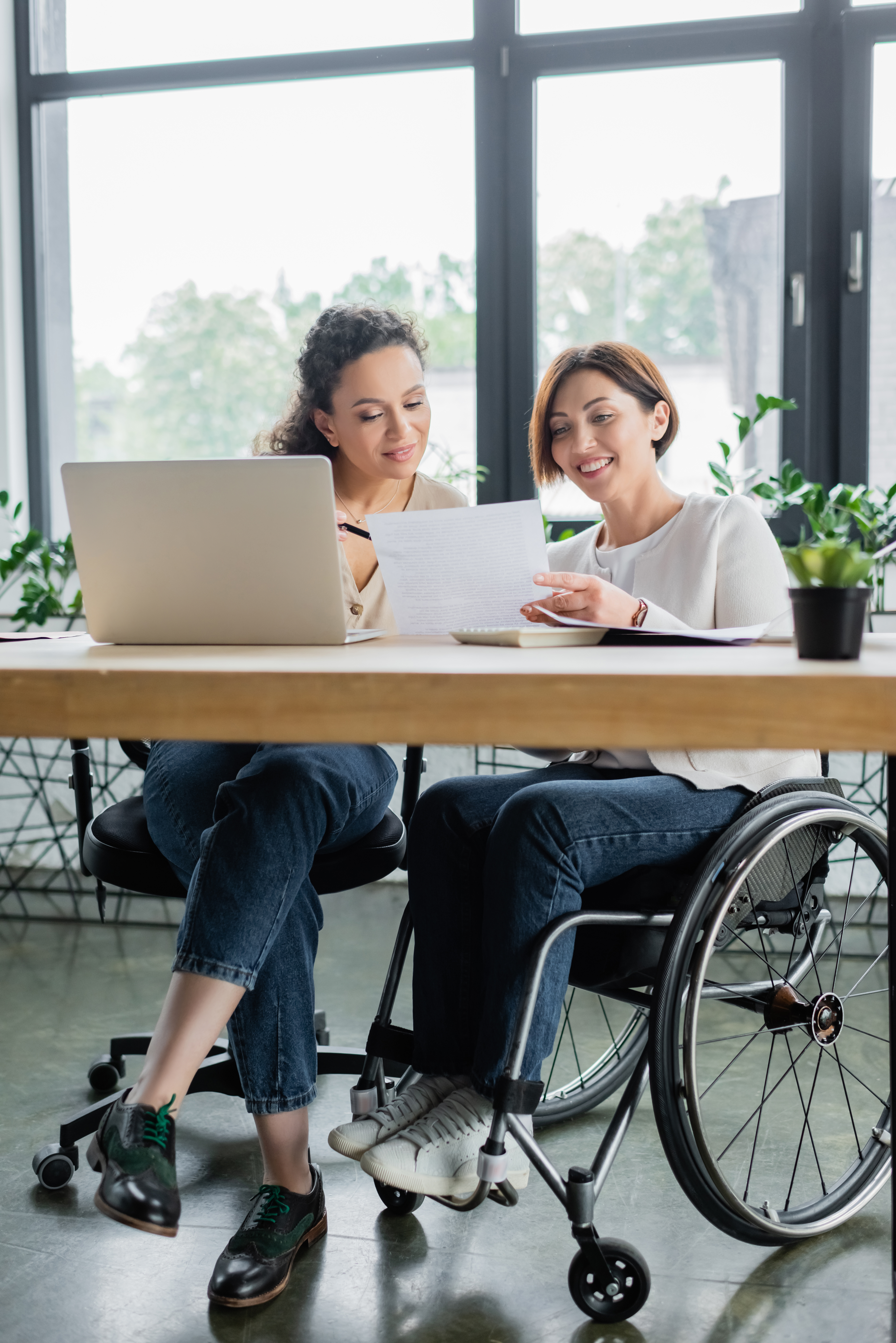 A woman sitting at a desk with a laptop and another woman in a wheelchair sitting showing her a paper.