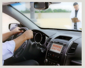 A driver’s arms hold a steering wheel in a car next to a display that says “warning: do not pass.” Outside of the car is a person holding a clipboard. 