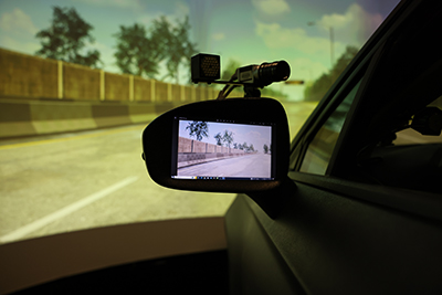 A closeup showing the side mirror of a car chassis in a driving simulator. The mirror is inlaid with a video screen and a camera on top of it. A wide, arching screen showing a virtual highway image surrounds the car.